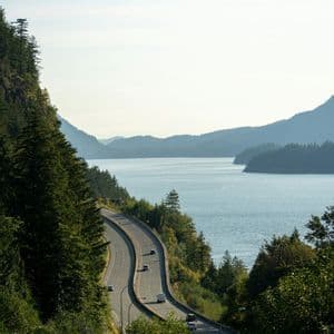 Una strada panoramica si snoda lungo la riva di un grande specchio d'acqua, incastonata tra ripide montagne ricoperte di alberi.