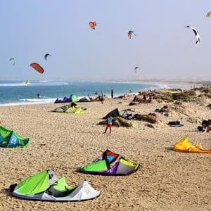 Una vasta spiaggia sabbiosa è animata da persone che fanno kitesurf, con molti aquiloni colorati sulla sabbia e nel cielo sopra l'oceano.