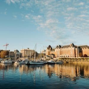 Una vista attraverso un porto calmo mostra barche a vela ormeggiate ed edifici della città riflessi nell'acqua sotto un cielo blu, parzialmente nuvoloso.