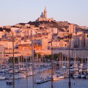 Sailboats docked in a harbor at sunset, with a city and a basilica on a hill in the background.