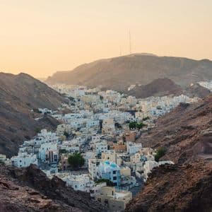 A town of white buildings nestled in a valley surrounded by rocky mountains at sunset.