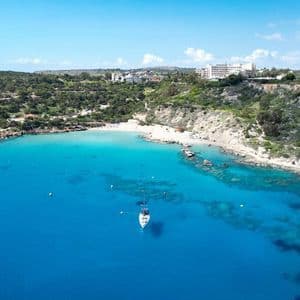 Una vista aerea di una barca a vela ancorata in una baia turchese accanto a una spiaggia sabbiosa appartata, con edifici sulle verdi colline sovrastanti.