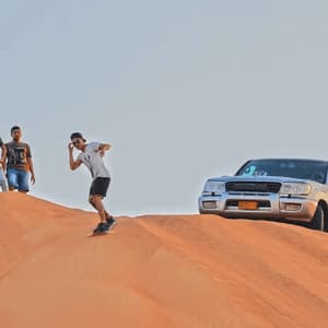 Ein WeRoad-Reisender sandboardet eine riesige Wüstendüne hinab, während seine Freunde und ein silberner SUV von oben zusehen.