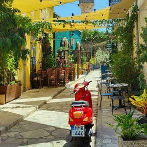 Uno scooter rosso parcheggiato in un vicolo acciottolato e illuminato dal sole, fiancheggiato da caffè colorati, tende da sole gialle e piante verdi, con un minareto sullo sfondo.