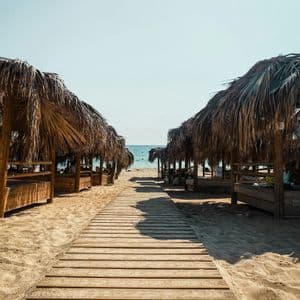 Una passerella di legno su una spiaggia sabbiosa conduce tra file di cabine con tetti di paglia verso l'oceano.