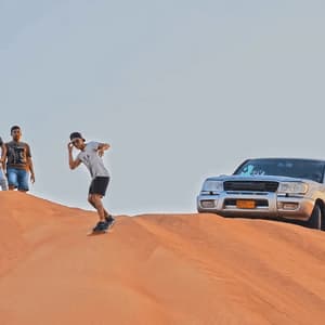 A person from a WeRoad group trip sandboards down an orange dune as others watch from the top next to a silver SUV.