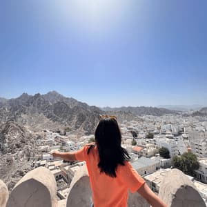 A woman in an orange shirt stands with arms outstretched, looking over a city nestled between mountains under a bright, sunny sky.