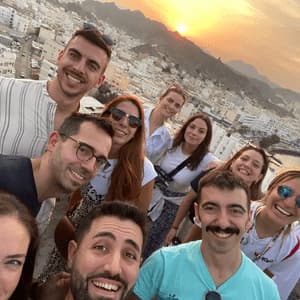 A WeRoad group trip takes a smiling selfie from a high viewpoint, overlooking a coastal city with white buildings at sunset.