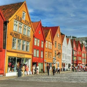 A row of colorful traditional wooden buildings with gabled roofs on a cobblestone street, with people walking in front.
