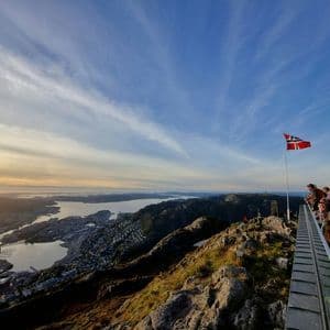 A Norwegian flag flies on a mountain viewpoint where people admire the panoramic view of a coastal city and bay at sunset.