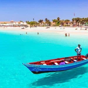 Un uomo è in piedi su una piccola barca blu che sembra galleggiare sull'acqua limpida e turchese vicino a una spiaggia sabbiosa.