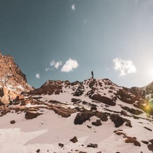 Eine Person steht auf einem schneebedeckten Berggipfel, als Silhouette vor strahlender Sonne und blauem Himmel.