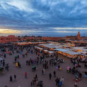 A high-angle view of a bustling city square at dusk, filled with crowds of people and rows of illuminated market stalls.