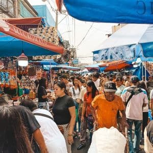 Una folla di persone riempie una strada stretta in un vivace mercato all'aperto, passando tra le bancarelle sotto tende blu e rosse.