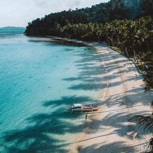 Una vista aerea di una spiaggia tropicale con una canoa a bilanciere in acqua turchese e lunghe ombre di palme sulla sabbia bianca.
