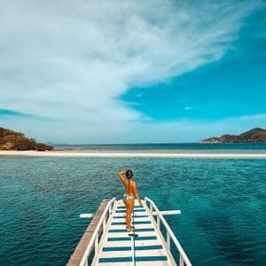 Una donna in bikini è a prua di una barca, che guarda una lingua di sabbia che collega due isole in un mare turchese.