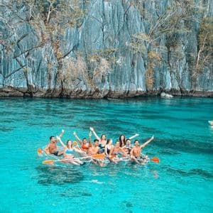 Un gruppo WeRoad sorridente e che saluta da kayak trasparenti su acqua cristallina turchese di fronte a una grande scogliera rocciosa.