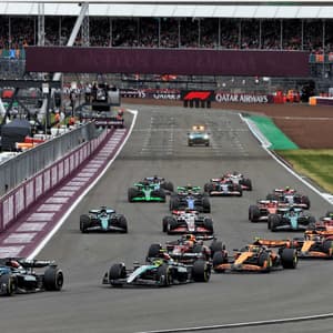 A pack of Formula 1 cars race down a track with grandstands full of spectators in the background.