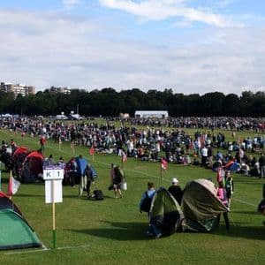 A large crowd of people gathers in a grassy field, setting up colorful tents for an outdoor event under a partly cloudy sky.