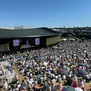 A large crowd of people sitting on a grassy hill watching a tennis match on a big screen at an outdoor venue on a sunny day.