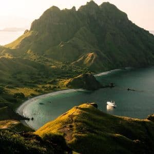 Una vista dall'alto di una barca bianca ancorata in una cala turchese, circondata da verdi montagne illuminate dal sole.