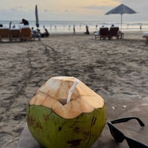 Un cocco verde con una cannuccia su un tavolo accanto a occhiali da sole su una spiaggia sabbiosa al tramonto.