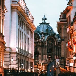 An ornate building with a large dome stands at the end of a cobblestone street where people are walking at dusk.