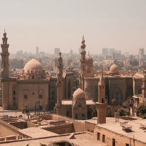 An aerial view of a large, sand-colored mosque with multiple domes and minarets against a hazy cityscape with modern skyscrapers in the background.