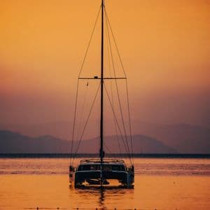 Un voilier catamaran ancré sur une eau calme, sous un ciel de coucher de soleil orange et des montagnes lointaines.