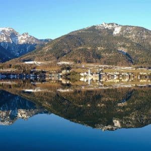 Un borgo lacustre e montagne innevate che si riflettono perfettamente nelle acque calme sotto un cielo azzurro.