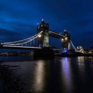 Eine große, beleuchtete Brücke mit zwei Türmen steht nachts über einem Fluss, ihre Lichter spiegeln sich auf der Wasseroberfläche wider.