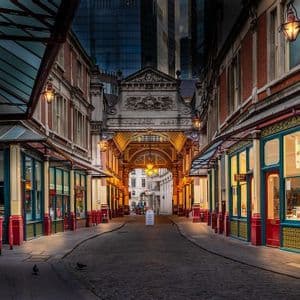 Eine leere, überdachte Kopfsteinpflastergasse mit verzierten, beleuchteten Ladenfronten, die in der Dämmerung zu einem großen Torbogen führt.