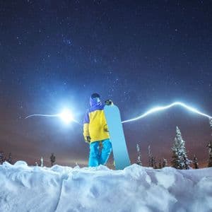 Uno snowboarder con giacca gialla tiene uno snowboard, in piedi su un paesaggio innevato sotto un cielo notturno stellato con scie luminose.