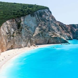 Veduta aerea di una spiaggia isolata di sabbia bianca e acqua turchese, ai piedi di una grande scogliera dalla cima verde, con un catamarano che naviga nelle vicinanze.