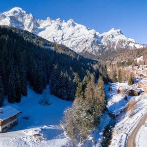 Una veduta aerea di un villaggio di montagna innevato immerso in una pineta, con grandi cime innevate sullo sfondo sotto un cielo azzurro.