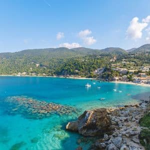 Una vista dall'alto di una baia costiera con acqua cristallina color turchese, una spiaggia sabbiosa e un piccolo villaggio immerso nelle verdi colline.