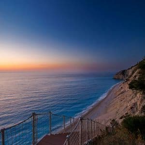 Una scala di legno con ringhiera in corda e rete scende da una scogliera verso una spiaggia sabbiosa e un mare turchese al tramonto.