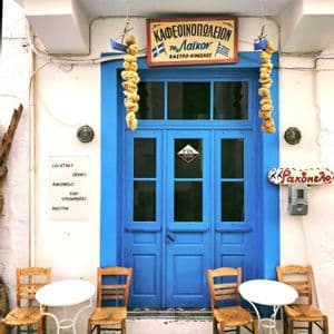 L'ingresso di un caffè in stile greco con una porta blu brillante, spugne marine appese e tavolini con sedie in legno all'esterno.