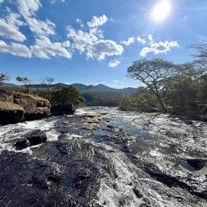 Un fiume ampio e roccioso scorre rapidamente sotto un sole splendente e un cielo azzurro nuvoloso, con colline boscose in lontananza.