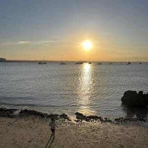 Un bambino piccolo cammina su una spiaggia sabbiosa al tramonto, con barche ancorate sull'acqua sullo sfondo.