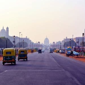 Two yellow and green auto-rickshaws driving on a wide road leading towards large domed buildings under a hazy sky.