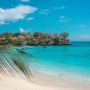 Una fronda di palma si protende su una spiaggia di sabbia bianca che costeggia una baia turchese, dove diverse piccole imbarcazioni sono ancorate vicino a una riva lussureggiante.