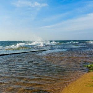 Onde dell'oceano si infrangono su una costa rocciosa e coperta di muschio, accanto a una spiaggia sabbiosa, sotto un cielo parzialmente nuvoloso.