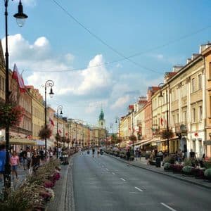 Una calle urbana larga y curva flanqueada por edificios históricos decorados con banderas polacas, con gente paseando por las aceras bajo un cielo azul.