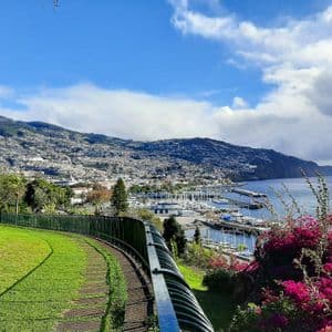 Ein Panoramablick von einer grünen Parkanlage auf eine Küstenstadt und einen Hafen, die am Fuße eines großen grünen Berges eingebettet sind.