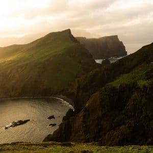 Eine Draufsicht auf steile, grün bewachsene Klippen, die zu einer abgelegenen Bucht im Meer unter einem goldenen, diesigen Himmel führen.