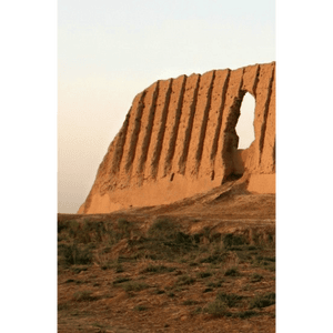 Una rovina di muro in mattoni di fango ondulato con una grande apertura si erge su una collina secca ed erbosa contro un cielo pallido e velato.