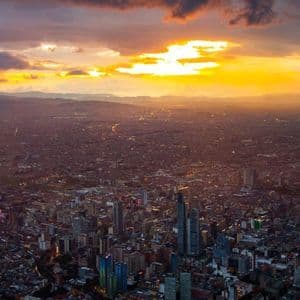 Una vista aerea di un'estesa città con edifici illuminati al tramonto, sotto un cielo drammatico con un brillante tramonto dorato.