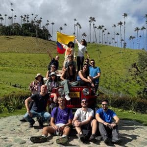 Un gruppo WeRoad in viaggio posa su e intorno a una jeep rossa in un paesaggio collinare con alte palme, con una persona che tiene una bandiera.