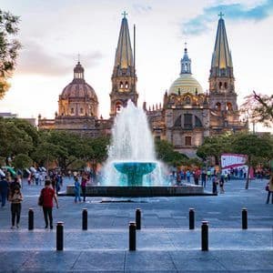 Una gran fuente de agua en una plaza de la ciudad con gente paseando frente a una majestuosa catedral con dos torres al atardecer.